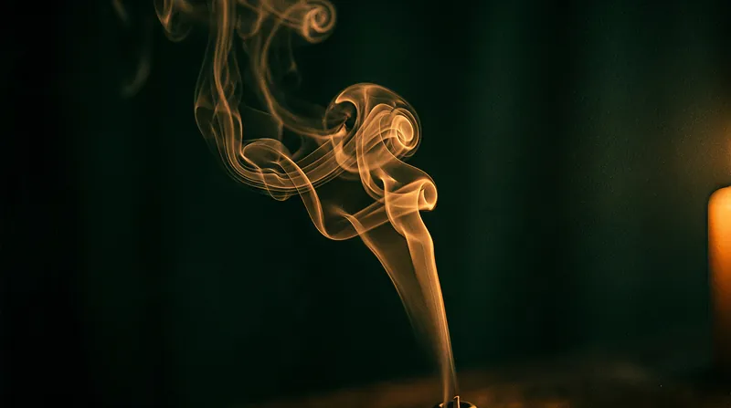 A thread of incense smoke rising in curls against a dark green background, with a blurred candle on the right